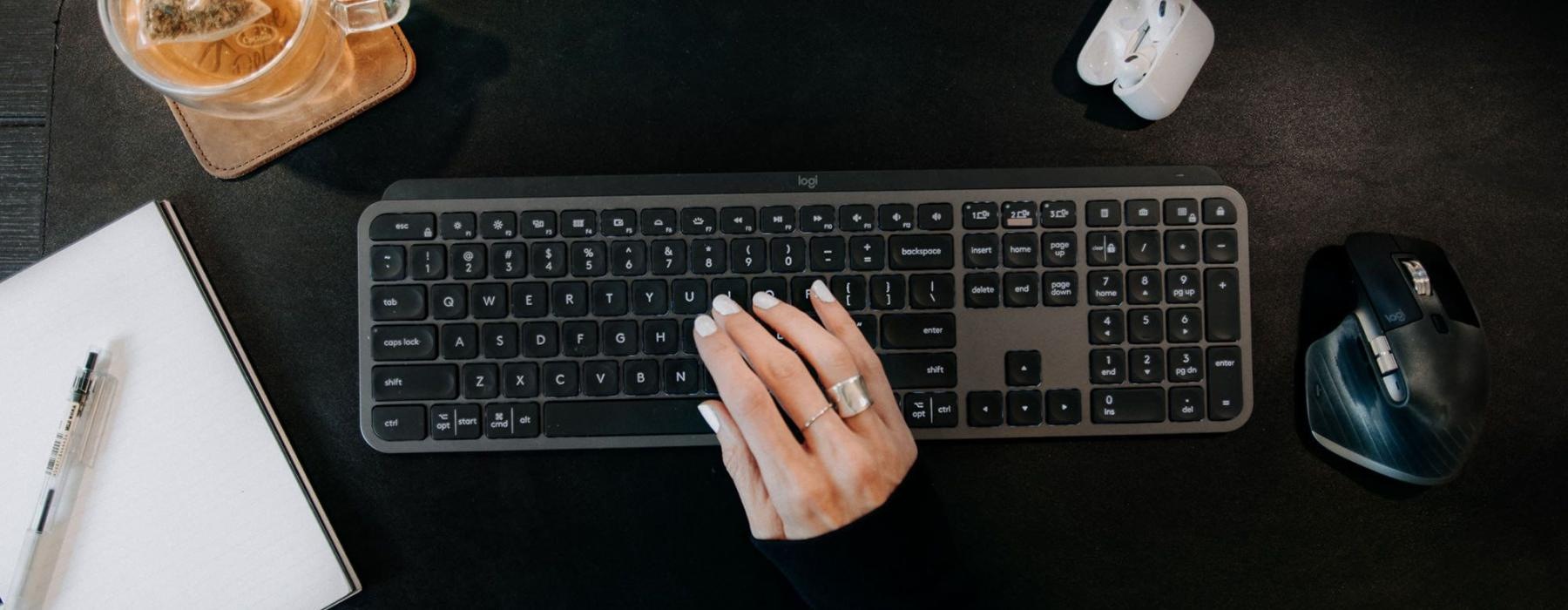 woman's hand on a keyboard surrounded by office items and a cup of tea on a coaster